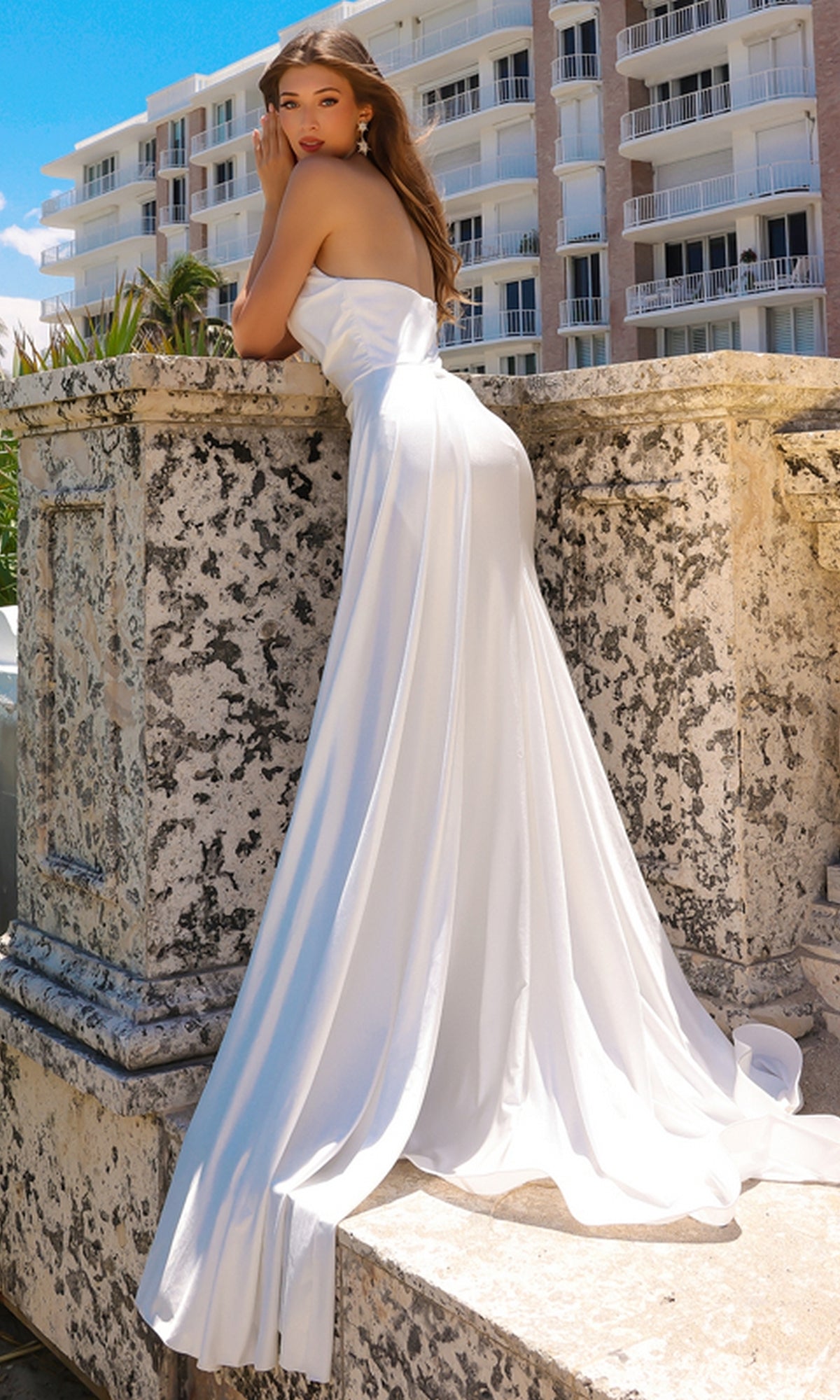 A woman in the Strapless Long Formal Prom Dress with Drape 3013 stands outdoors, leaning against a stone railing with a building and sky in the background.